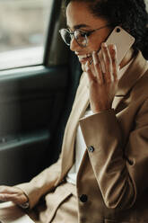 Female executive working on laptop and talking on cell phone while travelling by her car. Businesswoman sitting on back seat of car using laptop and phone. - JLPSF13194