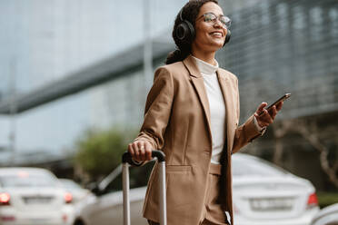 Female in businesswear ordering taxi online with her luggage while standing on a street. Smiling woman traveller with headphones standing on street with luggage bag listening to music on mobile phone. - JLPSF13199