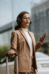 Businesswoman standing on the street with luggage and mobile phone. Female business traveller waiting for her taxi to arrive. - JLPSF13200