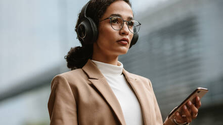 Confident female executive listening to music on cell phone. Businesswoman wearing headphones and holding a phone while standing outdoors on city street. - JLPSF13201