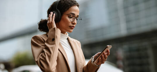 Businesswoman listening to music on her mobile phone. Businesswoman wearing headphones and looking at her cell phone on city street. - JLPSF13203