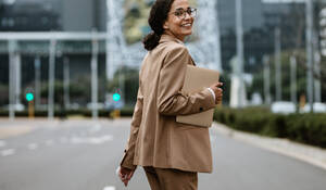 Businesswoman with a digital tablet walking on city street looking back at camera and smiling. Female business professional going home after work. - JLPSF13209