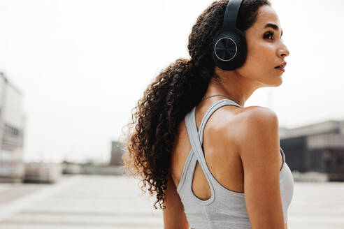 Fitness-Frau trägt Kopfhörer, um während des Trainings Musik zu hören. Frau in Sportkleidung macht Pause, steht im Freien und schaut weg. - JLPSF13221