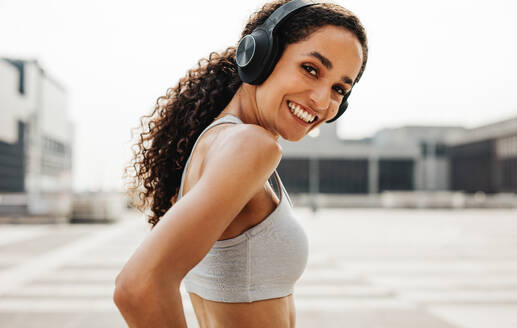Portrait of beautiful fitness woman wearing headphones looking at camera and smiling. Woman in sportswear exercising outdoors in the city. - JLPSF13223