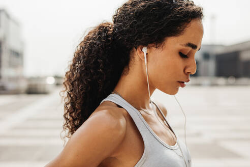 Sportswoman standing outdoors with earphones. Female athlete taking a break after intense training session. - JLPSF13225