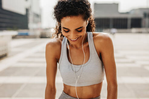 Fitness-Frau, die eine Trainingspause einlegt, nach unten schaut und lächelt. Frau in Sportkleidung hört Musik über Kopfhörer, während sie im Freien in der Stadt trainiert. - JLPSF13227