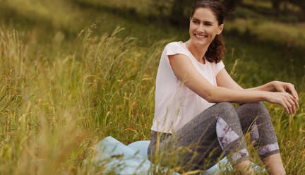 Lächelnde Frau, die in einem Park sitzt und wegschaut. Porträt einer fröhlichen Frau, die im Gras sitzt und die Natur genießt. - JLPSF13241