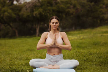 Frau sitzt auf einer Fitnessmatte in einem Park und übt Yoga. Porträt einer Frau, die in einem Park in einer Yoga-Pose sitzt. - JLPSF13243