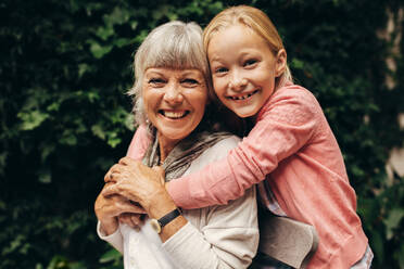 Nahaufnahme eines lächelnden Kindes, das seine Oma von hinten umarmt. Glückliche Großmutter und Kind in einem Park, die sich gegenseitig umarmen. - JLPSF13249