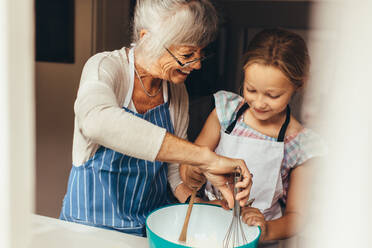 Ältere Frau bringt einem Kind bei, wie man Kuchenteig macht. Lächelnde Großmutter und Mädchen mischen Teig in einer Schüssel. - JLPSF13252