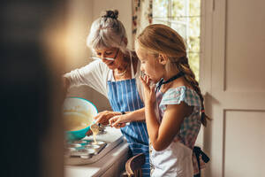 Großmutter bringt Kind bei, wie man Tassenkuchen macht. Glückliche Großmutter und Kind gießen Kuchenteig in Tassenkuchenformen. - JLPSF13257