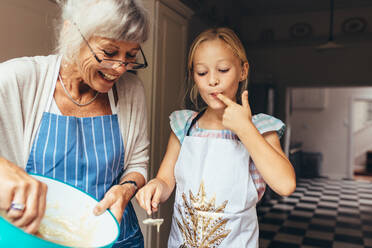 Ältere Frau in Schürze bei der Herstellung von Kuchenteig. Kleines Mädchen probiert mit Großmutter in der Küche stehenden Kuchenteig. - JLPSF13258