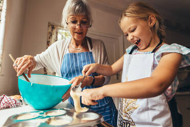 Kleines Mädchen lernt mit ihrer Großmutter, Tassenkuchen zu backen. Aufgeregtes Mädchen gießt Kuchenteig in Tassenkuchenformen. - JLPSF13259