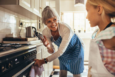 Lächelnde ältere Frau in Schürze öffnet die Ofentür. Glückliche Oma und Kind in der Küche beim gemeinsamen Kochen. - JLPSF13260