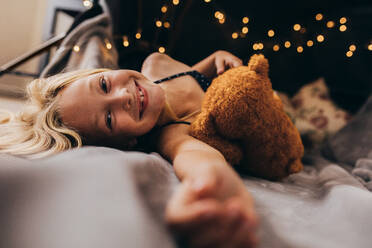 Smiling girl relaxing on bed holding her teddy. Close up of a happy kid lying on bed at home. - JLPSF13263