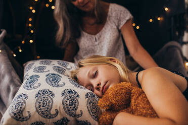 Kleines Mädchen schläft und hält einen Teddybär. Mädchen schläft auf dem Bett zu Hause mit ihrer Oma, die an ihrer Seite sitzt. - JLPSF13264