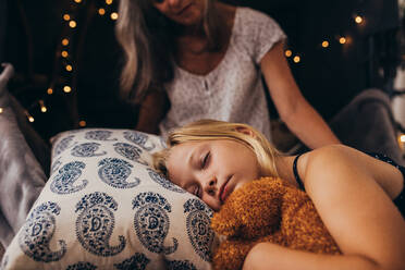 Little girl sleeping holding a teddy bear. Girl sleeping on bed at home with her granny sitting by her side. - JLPSF13264