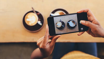 POV-Aufnahme einer Frau, die mit ihrem Smartphone Kaffeetassen auf dem Kaffeetisch fotografiert. Eine Frau fotografiert mit ihrem Mobiltelefon Lebensmittel für ihre sozialen Medien. - JLPSF13273