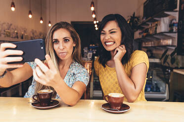 Frau streckt ihre Zunge heraus, während sie ein Selfie mit einem lächelnden Freund im Café macht. Lustige Freundinnen, die ein Selfie mit dem Handy im Café machen. - JLPSF13276