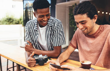 Afrikanischer Mann mit einer Kamera im Gespräch mit einem Freund, der sein Smartphone in einem Café benutzt. Kreative Menschen treffen sich in einem Café. - JLPSF13290