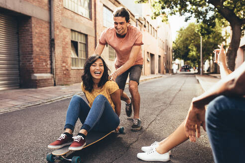 Frau, die auf einem Skateboard sitzt und von einem männlichen Freund geschoben wird, während andere Freunde, die daneben sitzen, ihre Bilder mit dem Smartphone besprechen. Freunde, die Spaß mit dem Skateboard auf der Straße haben. - JLPSF13291