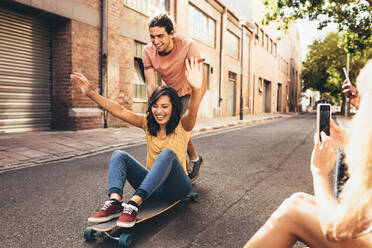 Paar hat Spaß auf dem Skateboard mit Freunden, die ihre Fotos machen. Mann schiebt Frau auf dem Skateboard mit ihren Freunden, die an der Straße sitzen und ihre Fotos mit dem Mobiltelefon machen. - JLPSF13292