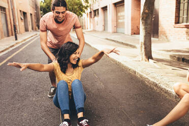 Excited young woman being pushing on skateboard by her boyfriend outdoors on street, with friends sitting by the street. Couple enjoying themselves outdoors. - JLPSF13295