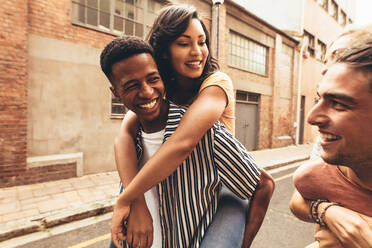 Two men giving piggyback ride to their girlfriends outdoors. Multi-ethnic friends having fun outdoors on city street. - JLPSF13303