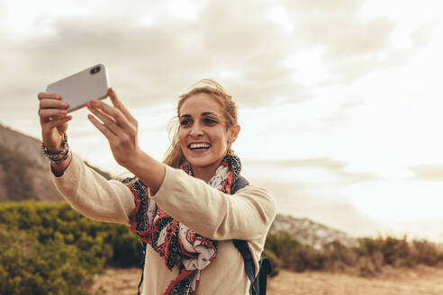 Lächelnde junge Frau, die ein Selfie mit ihrem Smartphone macht, während sie auf einem Berg wandert. Eine Wanderin macht ein Selfie für ihre sozialen Medien. - JLPSF13309