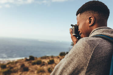 Afrikanischer Mann, der mit seiner Digitalkamera Bilder von einer schönen Meereslandschaft macht und neue Inhalte für seine sozialen Medien aufnimmt. - JLPSF13313