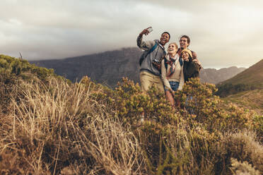 Eine Gruppe von Kreativen auf einem Wanderausflug macht ein Selfie, um neue Inhalte für soziale Medien zu erstellen. Junge Männer und Frauen wandern gemeinsam auf einem Bergpfad, während ein männlicher Freund ein Selfie mit seinem Smartphone macht. - JLPSF13321