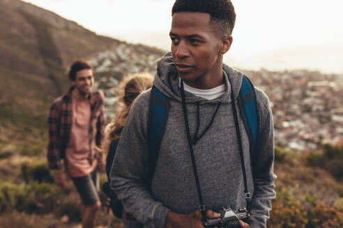 Afrikanischer Mann mit Kamera beim Wandern auf einem Bergpfad mit seinen Freunden im Hintergrund. Junge Leute auf Wandertour. - JLPSF13322