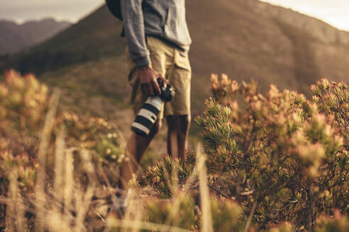 Mittelaufnahme eines Mannes, der mit seiner DSLR-Kamera auf einem Bergpfad steht, Fokus auf Wildpflanzen auf dem Bergpfad mit einem Fotografen im Hintergrund. - JLPSF13326