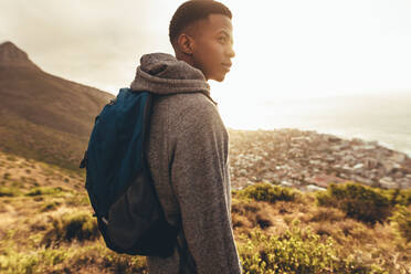 Junger afrikanischer Mann mit Rucksack, der auf einem Hügel steht und eine Aussicht mit hellem Sonnenlicht von hinten betrachtet. Junger Mann auf Wandertour. - JLPSF13327