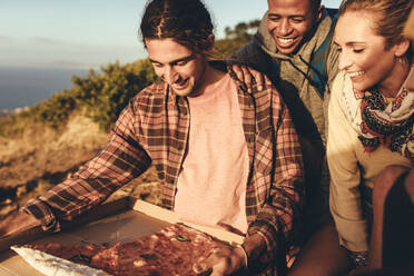 Eine Gruppe von Wanderern isst Pizza. Ein Mann mit Pizza entspannt sich auf einem Bergpfad, während seine Freunde daneben sitzen. - JLPSF13332