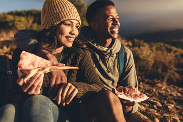 Junger Mann und Frau sitzen auf einem Bergpfad, essen Pizza und genießen die Aussicht. Paar auf Wandertour isst Pizza und bewundert die Aussicht. - JLPSF13335