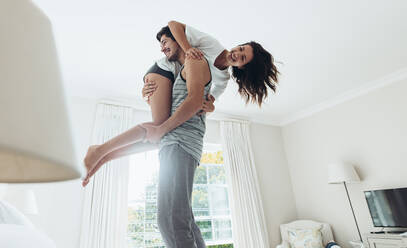 Young man standing on bed and carrying his girlfriend on his shoulder. Couple in playful mood in bedroom. - JLPSF13357