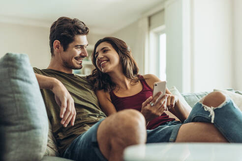Verliebtes Paar sitzt zu Hause auf dem Sofa. Mann und Frau entspannen sich auf der Couch mit Smartphone im Wohnzimmer. - JLPSF13361