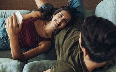 Junge Frau mit Smartphone auf dem Schoß ihres Freundes liegend und lächelnd. Paar entspannt sich auf dem Sofa zu Hause. - JLPSF13363