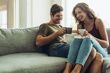 Happy young couple relaxing on couch at home with coffee. Young man and woman relaxing on sofa drinking coffee - JLPSF13364