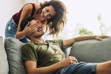 Young woman standing behind her boyfriend on sofa and covering his eyes at home. Woman surprising man at home. - JLPSF13366