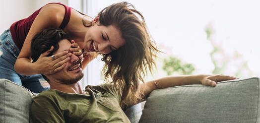 Lächelnde junge Frau, die ihrem Partner auf dem Sofa sitzend die Augen zuhält. Junge Frau überrascht Mann im Wohnzimmer zu Hause. - JLPSF13367