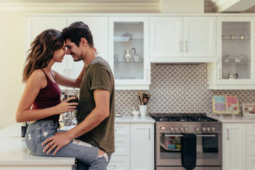 Attractive passionate couple in kitchen. Woman is sitting on kitchen counter with her boyfriend standing by, both touching their noses. - JLPSF13375