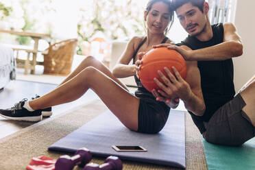 Gesundes junges Paar, das zu Hause Übungen mit einem Ball macht. Das Paar sitzt Rücken an Rücken auf einer Yogamatte und übt, indem es sich gegenseitig den Ball zuspielt. - JLPSF13387