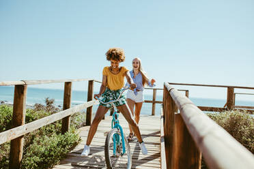 Fröhliche Frau auf dem Fahrrad auf der Promenade mit einem Freund, der hinter ihr läuft. Weibliche Freunde genießen ihren Urlaub. - JLPSF13388
