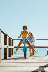 Woman pushing her friends sitting on a bicycle. Two girls enjoying on their vacation at the beach. - JLPSF13390