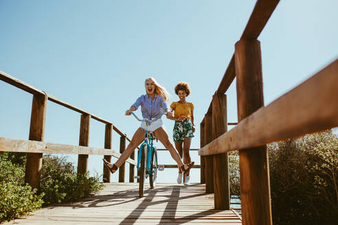 Eine aufgeregte Frau fährt mit dem Fahrrad die Promenade hinunter, während ihre Freunde hinter ihr herlaufen. Zwei junge Freundinnen haben eine tolle Zeit in ihrem Urlaub. - JLPSF13393