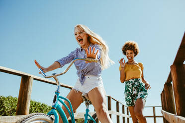 Excited girl cycling on a boardwalk with her friends running. Two woman friends enjoying themselves on the vacation. - JLPSF13394