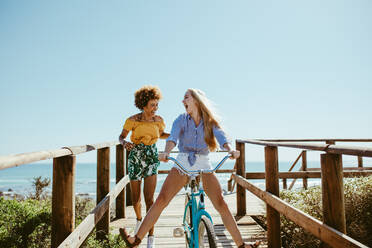 Young woman riding bike on boardwalk with friend running by. Multi-ethnic girls having fun with a bike at the seaside boardwalk. - JLPSF13395