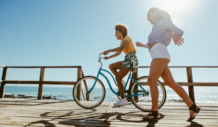 Junge Frau auf dem Fahrrad mit ihrer Freundin, die an der Strandpromenade vorbeifährt. Multi-ethnische Freundinnen, die sich an der Strandpromenade mit dem Fahrrad vergnügen. - JLPSF13404